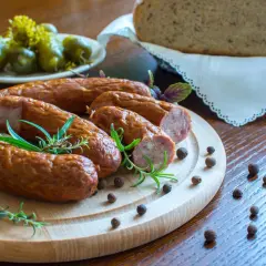 Kiełbasa na desce z rozmarynem, obok ogórki, chleb i ziarna pieprzu.