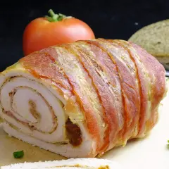 Rolada z indyka w boczku, idealna do krojenia bez rozpadania się. Na desce z boczku, obok pomidora i chleba.