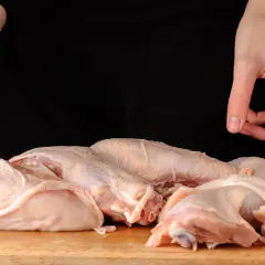 Surowe kawałki kurczaka na desce, ręce trzymające nóż i palec wskazujący.