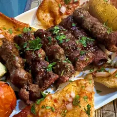 Talerz z grillowanymi szaszłykami jagnięciny, pieczonymi ziemniakami, pomidorem i chlebem.