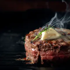 Soczysty stek wołowy z masłem i rozmarynem na grillu, z którego unosi się para.