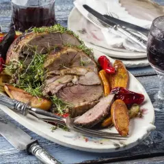 Pieczona jagnięcina z warzywami i ziołami na talerzu, obok kieliszek wina.
