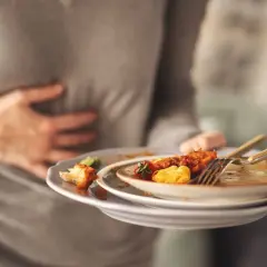 Osoba trzymająca brudne talerze po jedzeniu, masująca bolący brzuch. Dlaczego po zjedzeniu wieprzowiny boli brzuch? Najczęstsze przyczyny.