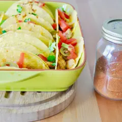 Domowa przyprawa do taco w słoiku obok talerza pełnego tacos z pomidorami i jalapeno. Lepsza niż gotowa z torebki!