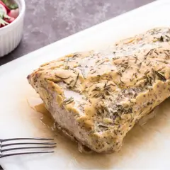 Marynata z maślanki do schabu sprawia, że mięso wieprzowe staje się delikatne i soczyste. Na desce leży pieczony schab z ziołami, obok miseczka z rzodkiewką i natką.