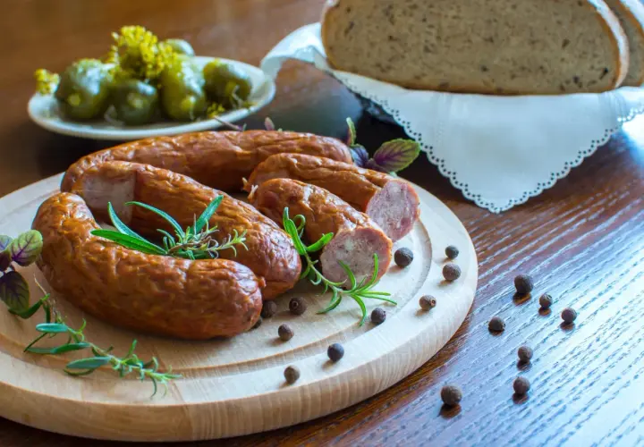 Kiełbasa na desce z rozmarynem, obok ogórki, chleb i ziarna pieprzu.