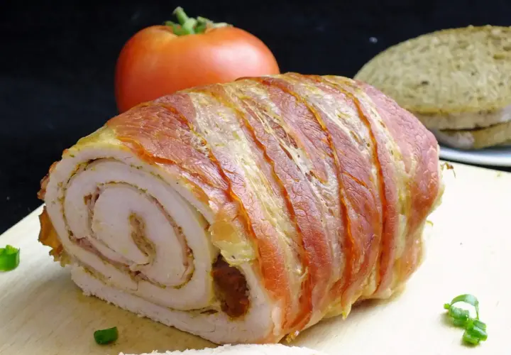 Rolada z indyka w boczku, idealna do krojenia bez rozpadania się. Na desce z boczku, obok pomidora i chleba.
