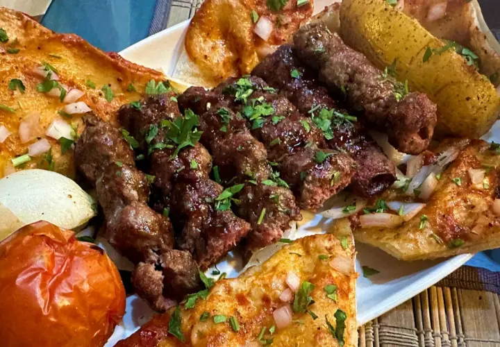 Talerz z grillowanymi szaszłykami jagnięciny, pieczonymi ziemniakami, pomidorem i chlebem.