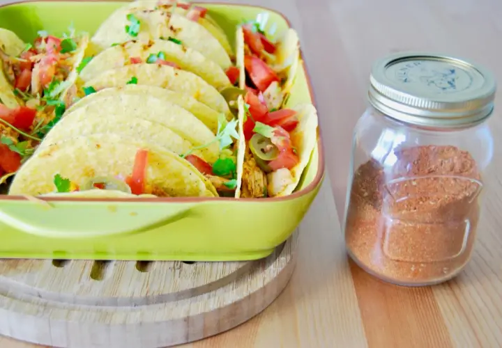 Domowa przyprawa do taco w słoiku obok talerza pełnego tacos z pomidorami i jalapeno. Lepsza niż gotowa z torebki!