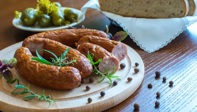 Kiełbasa na desce z rozmarynem, obok ogórki, chleb i ziarna pieprzu.