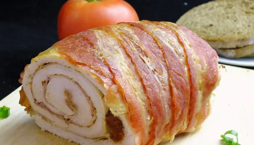 Rolada z indyka w boczku, idealna do krojenia bez rozpadania się. Na desce z boczku, obok pomidora i chleba.