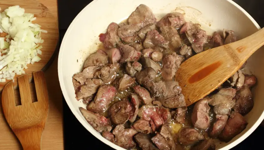 Smażona wątróbka w patelni z drewnianą łopatką, obok posiekana cebula.