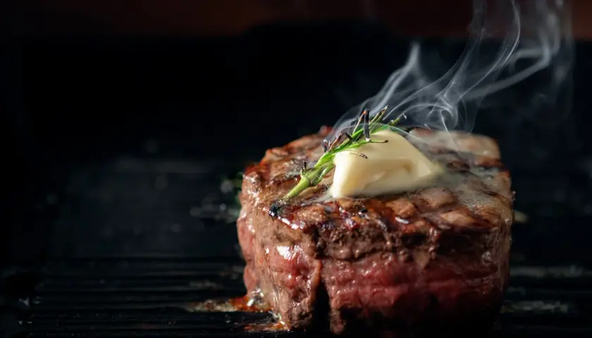 Soczysty stek wołowy z masłem i rozmarynem na grillu, z którego unosi się para.