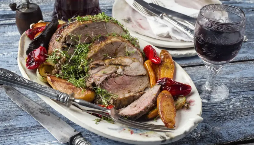Pieczona jagnięcina z warzywami i ziołami na talerzu, obok kieliszek wina.
