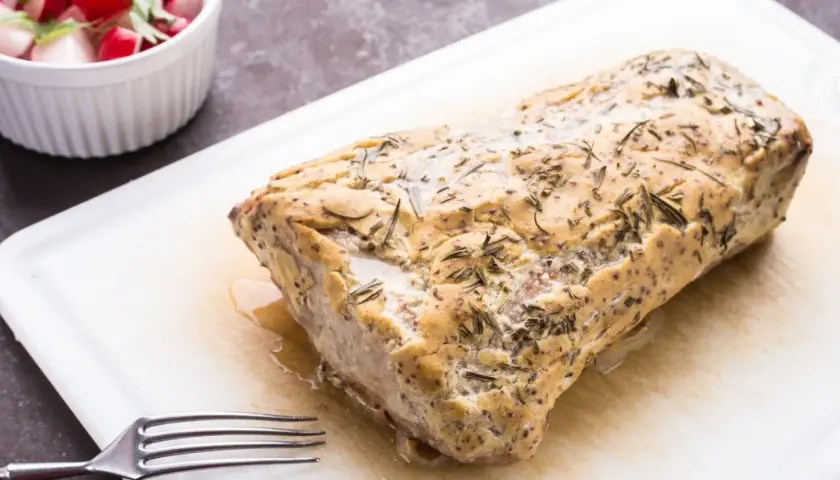 Marynata z maślanki do schabu sprawia, że mięso wieprzowe staje się delikatne i soczyste. Na desce leży pieczony schab z ziołami, obok miseczka z rzodkiewką i natką.