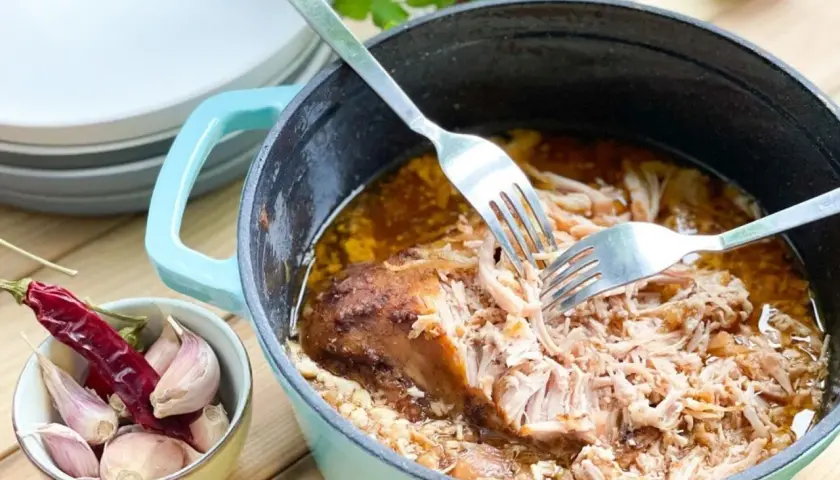 Szarpana wieprzowina w sosie, rozrywana widelcami w naczyniu żeliwnym, obok czosnek i papryczka chili.