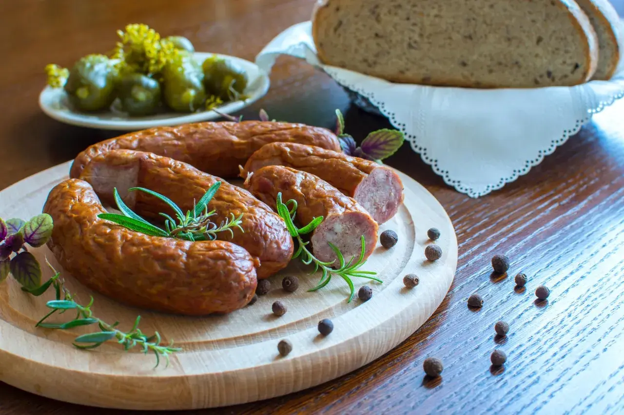 Kiełbasa na desce z rozmarynem, obok ogórki, chleb i ziarna pieprzu.