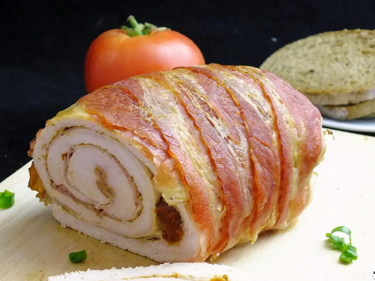 Rolada z indyka w boczku, idealna do krojenia bez rozpadania się. Na desce z boczku, obok pomidora i chleba.