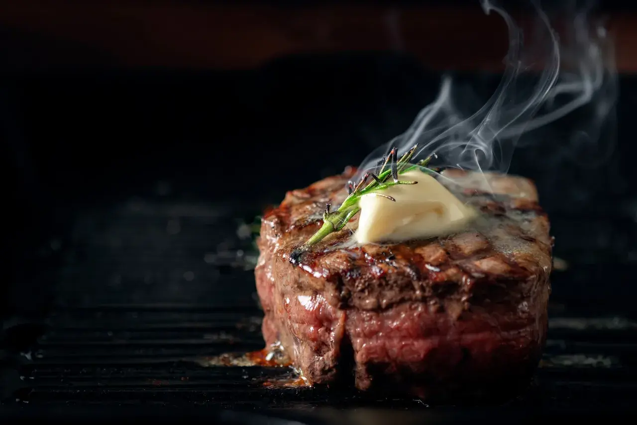 Soczysty stek wołowy z masłem i rozmarynem na grillu, z którego unosi się para.