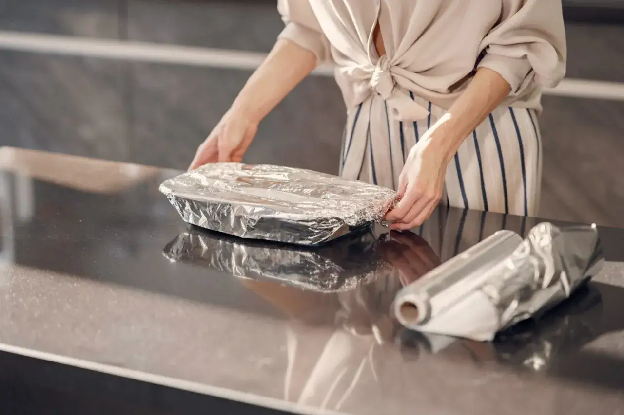Przygotowanie do pieczenia mięsa w folii aluminiowej. Czy błyszcząca strona ma znaczenie? Kobieta owija naczynie folią.